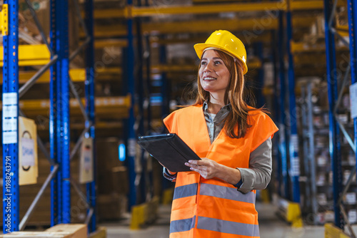 Female Warehouse Worker Wearing Hard Hat And High Visibility Vest With Tablet Inspecting Inventory