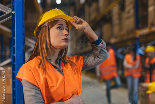 Female Warehouse Worker in Hard Hat and Orange Safety Vest Inspecting Inventory in Industrial Warehouse