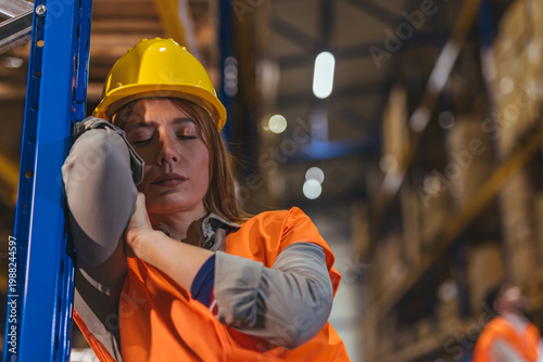 Tired Female Warehouse Worker Wearing Hard Hat and High Visibility Vest Resting During Night Shift