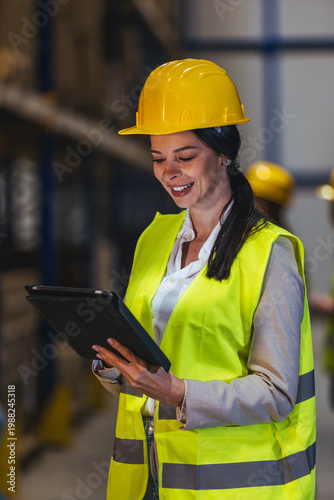 Female Warehouse Manager Wearing Hard Hat And Safety Vest Using Tablet For Inventory And Logistics