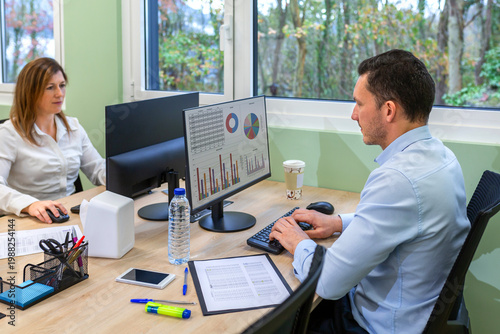 Business colleagues analyzing data and working in a coworking office. Concentrated young businessman typing on computer keyboard while checking documents and charts for financial analysis project.
