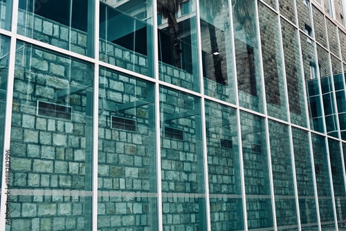 Modern architecture building facade featuring glass windows reflecting the sky and a textured stone wall background.