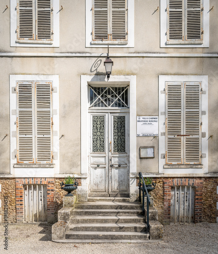 Charming rustic French countryside house façade featuring weathered wooden shutters, aged double doors with decorative wrought iron, and a small stone staircase leading to the entrance. 
