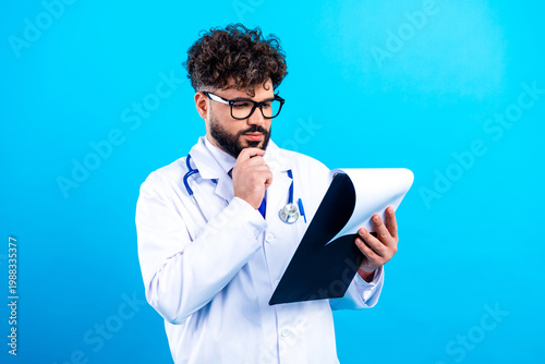 Tablou pe pânză Male doctor in white coat with stethoscope reading clipboard and thinking agains