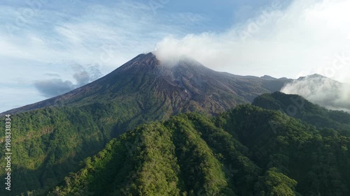 An aerial view of Mount Merapi surrounded by dense forest in Java, Indonesia. A view of volcanic terrain and hills under a clear blue sky.
