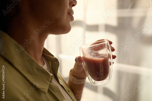 Foto Close up of young adult woman holding glass mug and drinking smoothie near window, suggesting morning wellness routine