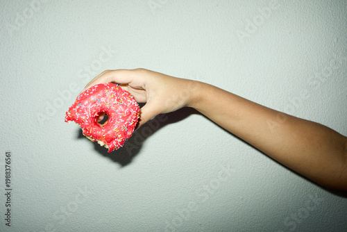 Fotografie Close up of adult hand holding bitten pink frosted donut with sprinkles against plain wall, suggesting indulgence and craving