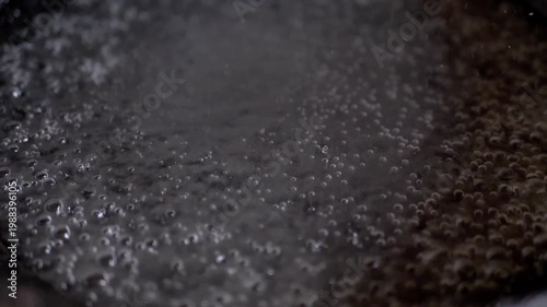 An intense close-up shows water rapidly boiling in a metallic pot, with steam rising and countless small bubbles forming and bursting. Captures the energy of heating liquid.