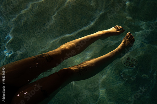 Fotografie Close-up of a person's legs and feet partially submerged in blue pool water with atmospheric low light and reflections