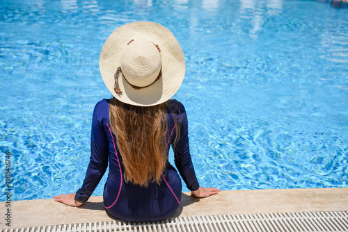 Woman in straw hat and long sleeve swimsuit sitting by swimming pool. Summer vacation at luxury resort, relaxation, skin protection from UV rays and wellness concept. Back view of female tourist
