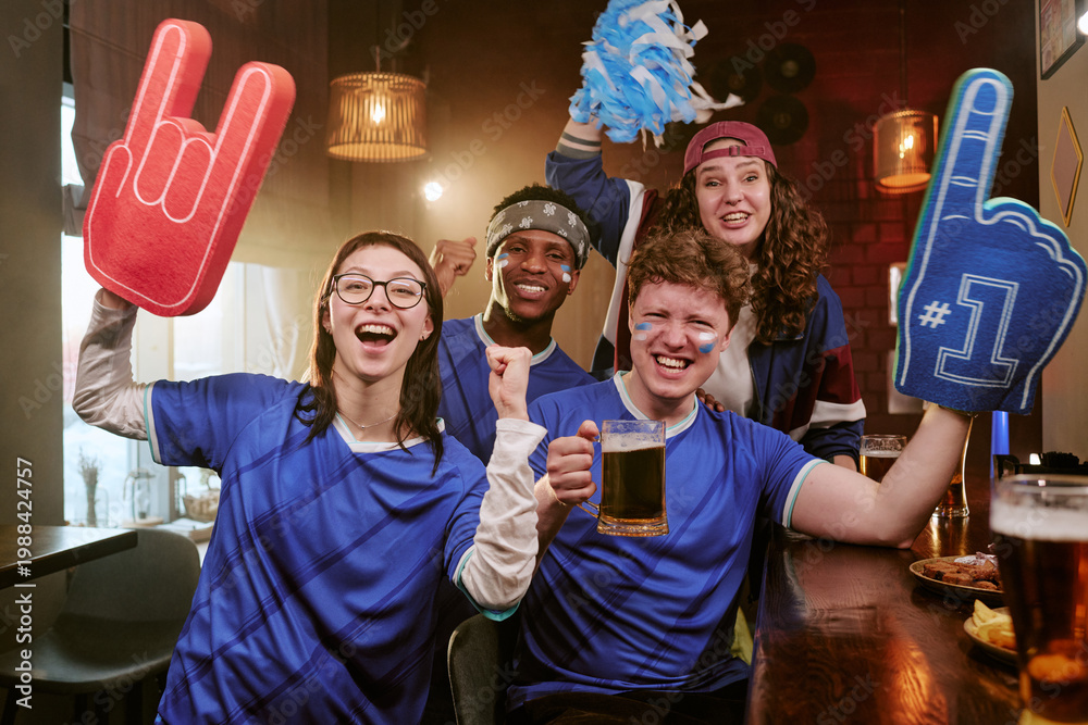 Fototapeta premium Portrait of diverse young adult sports fans cheering in bar, raising foam fingers and beer mugs during match viewing. Ideal for fan culture, team support, sports marketing campaigns