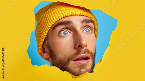 Bright yellow background with torn paper effect. Young man peers through a hole, eyes wide, mouth open. Beard and knit cap add playful contrast. Surprise or discovery conveyed with bold colors