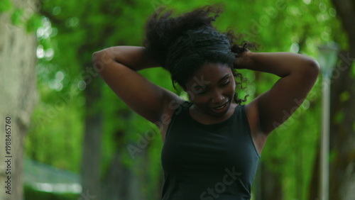 African American woman standing outdoors adjusting her hair with both hands creating natural and candid personal care moment in green park environment