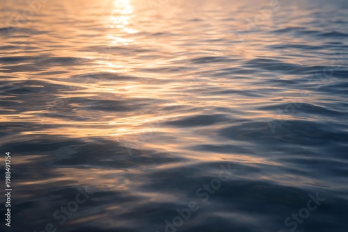 Fotografia Close-up of rippling water reflecting the warm glow of the setting sun, creating a serene and tranquil natural scene with gentle waves and shimmering light