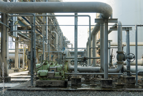 Large industrial piping network with valves and support structures. Metal pipes run horizontally and vertically across the scene. Concrete bases and grates form the ground