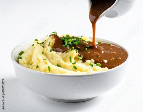 Delicious creamy mashed potatoes topped with rich brown gravy and fresh green chives being poured from a white gravy boat in a studio shot on a clean white background