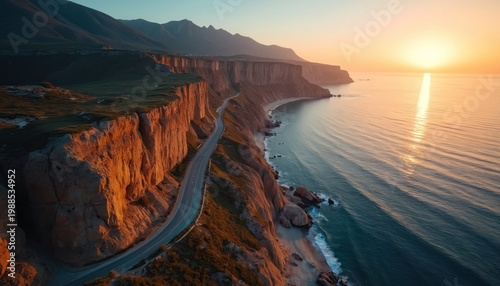 Curvy road runs along steep coastal cliffs beside calm ocean at golden sunset. Mountains rise in distance under warm sky light. Scenic natural landscape.