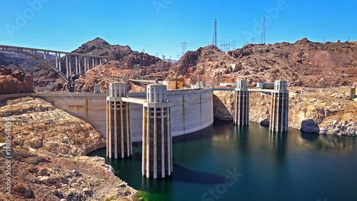 Hoover Dam and bridge in desert canyon. Wide view of Hoover Dam with reservoir towers, arched bridge, rocky desert mountains, and clear blue sky in the American Southwest.