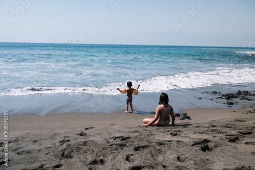 A sun-drenched beach with gentle waves breaking on the shore. Children are playing near the water’s edge. Clear skies, calm seas, and a warm, relaxed atmosphere.