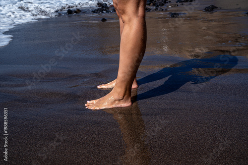 Feet step gently on dark, wet sand near ocean edge. Waves roll softly in the background, creating misty foam. Shadow stretches long on damp ground beside bare legs. Scene captures quiet