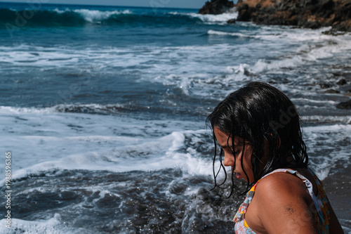 Young girl stands at ocean's edge, water splashing. Dark hair clings to her face as waves crash close. Sunlight glints off foam, casting soft shadows. She gazes at sea, quiet