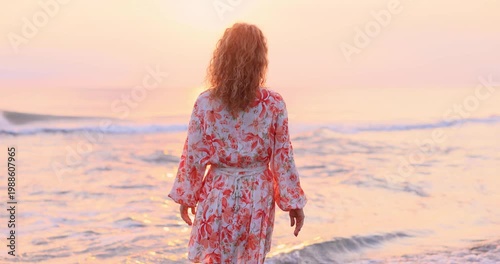 Happy woman is walking on the sandy beach in a red dress against sunrise. Slow motion