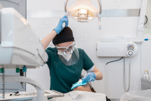 Female dentist in protective gear adjusting medical lamp while performing tooth treatment with dental dam. Professional healthcare service for oral hygiene, stomatology and surgery process.