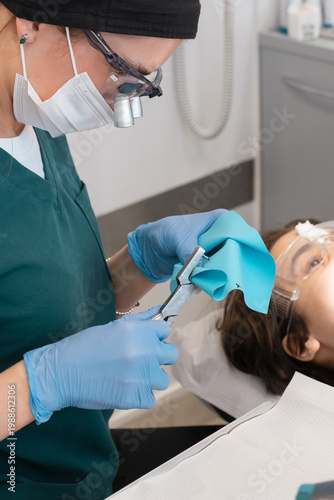 Rubber dam punch tool used by professional dentist to prepare blue latex sheet for patient treatment. Dental isolation equipment for tooth restoration in clinic office with specialist.