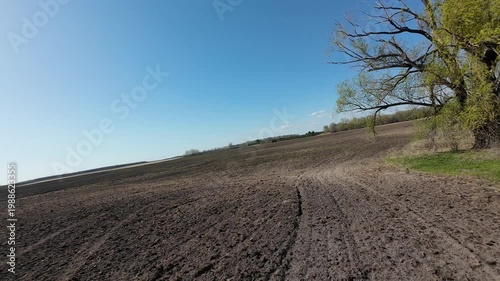Wide angle FPV drone shot flying fast over dark plowed soil in countryside. Panoramic view of agricultural land with green meadow and lone tree under bright sun during spring day.