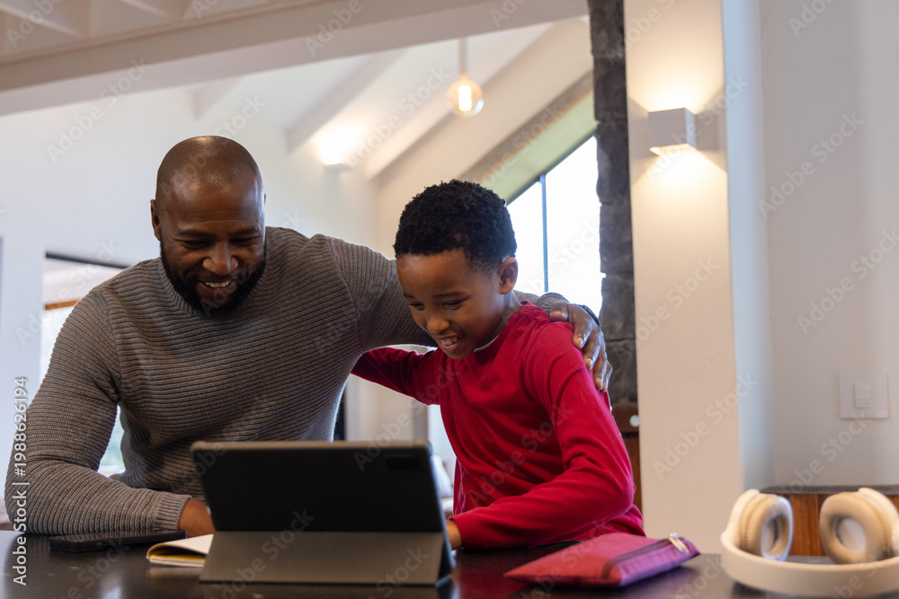 Obraz premium African American father and school-age son leaning over tablet at kitchen counter, smiling together