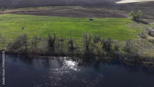 High angle aerial shot of dark car parked on green meadow near river with bright sun reflection. Picturesque countryside landscape featuring blue water, spring trees and plowed agricultural field.