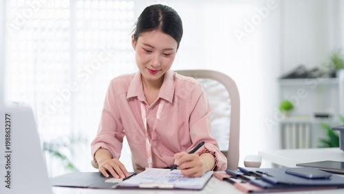 Obraz na płótnie Focused Work: An Asian woman diligently engrossed in work, carefully reviewing documents while seated at her desk in a well-lit office