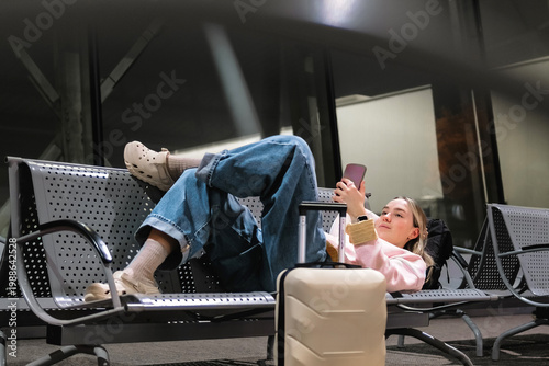 Photos Young female traveler resting on airport bench with suitcase and smartphone, delayed flight and layover concept, night travel, waiting for departure and modern solo travel lifestyle