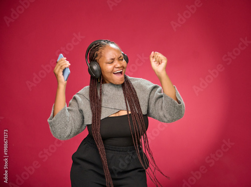 Young Black woman with long braids joyfully singing and dancing while wearing over-ear headphones and holding smartphone against vibrant pink studio background, conveying energy and happiness