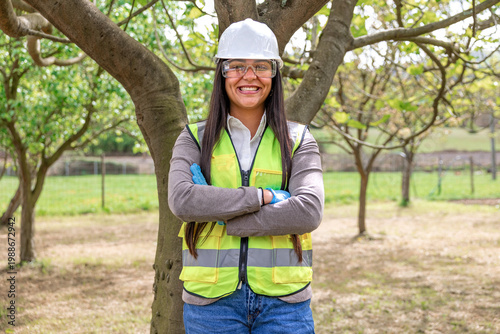 Latin woman engineer wearing hard hat, safety vest, and protective glasses, smiling with arms crossed outdoors, representing environmental protection and sustainable development