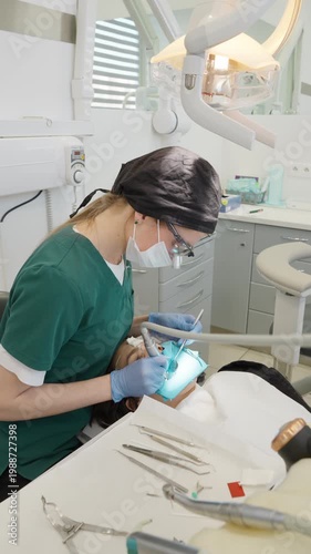 Professional dentist performs tooth cavity preparation using dental drill. Assistant uses suction. Female doctor treats patient with blue rubber dam in medical clinic office procedure.