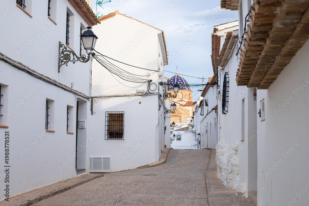 Fototapeta premium Spain, Altea city. Narrow cobblestone street lined with whitewashed buildings and traditional architecture in a quaint village setting under a clear sky