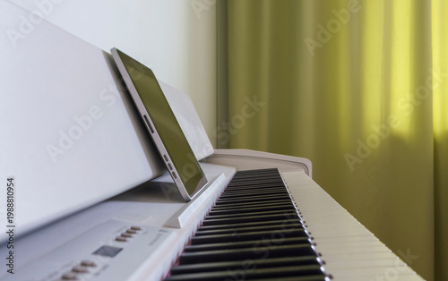 Digital piano with black and white keys positioned beside a tablet on a white surface, with green curtains softly diffusing light in the background
