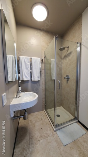 Modern bathroom with glass shower stall and sink. White towels hang neatly on a rack. Compact space features tiled floors and walls. Round ceiling light illuminates the room evenly