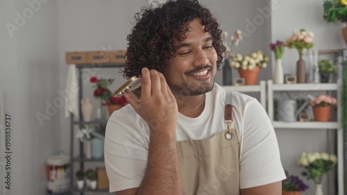Man florist holding smartphone to ear and smiling in a studio shop wearing apron; small business joy.