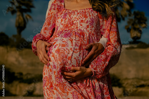Pregnant woman in beautiful long dress with husband on sand near ocean. Mother and father hugging while expecting baby on sunny day