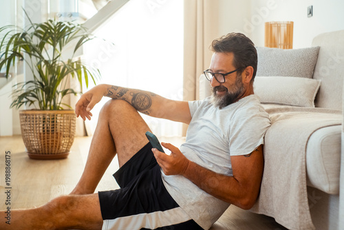 Schilderij op canvas Relaxed middle-aged man with tattoo using smartphone while leaning against sofa