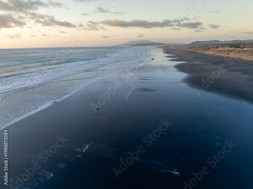 Fotografie A lone figure walks along a wet, black sand Kawhia Hot Water Beach, Kawhia, Waikato, New Zealand, with waves gently lapping the shore and dunes in the background