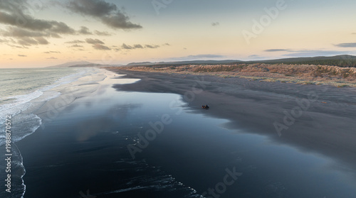 Canvas Print A lone ATV drives along a vast black sand Kawhia Hot Water Beach, Kawhia, Waikato, New Zealand, with gentle waves lapping the shore and dunes in the background