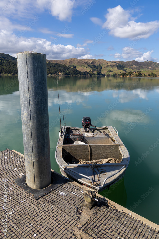 Fototapeta premium A small fishing boat with an outboard motor is moored to a jetty on a calm, reflective bay in Kawhia Harbour, Te Waitere, Waikato, New Zealand. The boat is ready for a day of fishing.