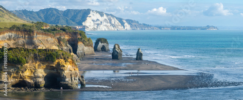 Fotografi Two people walk along the beach at the The Three Sisters, Tongaporutu, New Zealand, with its iconic rock formations and sea stacks