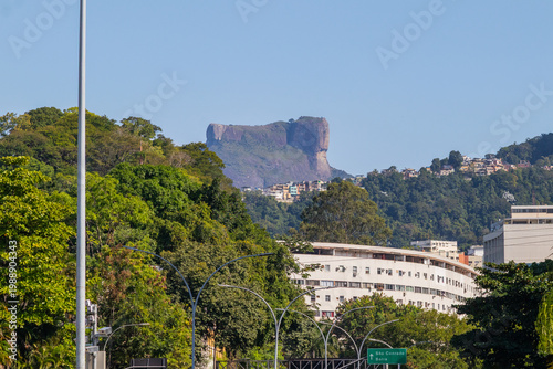 Scenic view of the iconic Pedra da Gávea mountain towering over the urban landscape and lush green forests of Rio de Janeiro, Brazil, featuring residential buildings under a clear blue sky