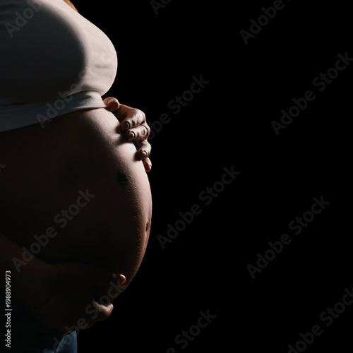 Close-up of pregnant belly held by woman hands in dramatic low key lighting, isolated on dark background, intimate and emotional mood, concept of motherhood, prenatal care and maternity