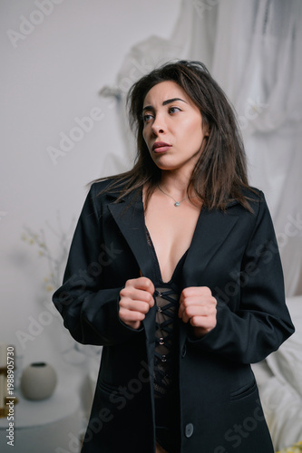 Elegant woman in black blazer and lace lingerie holding lapels in minimalist studio with soft drapery, natural light and neutral tones, sensual fashion portrait for branding, editorial and advertising