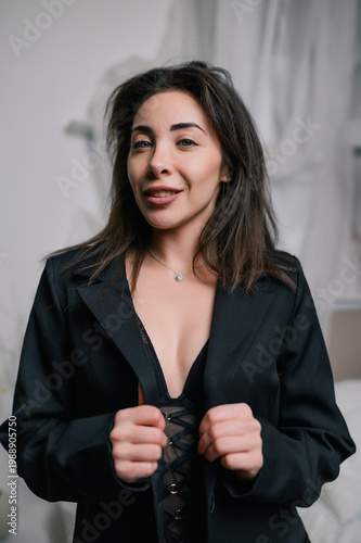 Smiling woman in black blazer and lace lingerie holding lapels in soft light studio with neutral tones and drapery, elegant sensual portrait for fashion, branding and advertising use